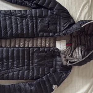 Svart Moncler Clovis Giubbotto 2017 dunjacka med huva - Säljer en svart dunjacka från Moncler med huva och grått foder. Jackan har två bröstfickor med lock, sidofickor med dragkedja och Moncler-logga på ärmen. Quiltad design och dubbel dragkedja framtill. Perfekt för kalla dagar. Pris kan diskuteras