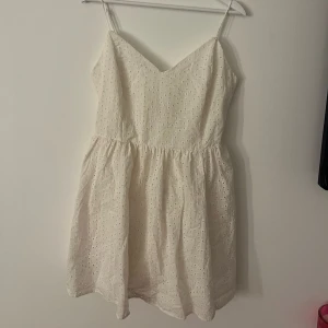 Vit somrig klänning med broderade detaljer - Söt vit kortklänning med tunna axelband och v-ringning. Klänningen har broderade hålmönster över hela tyget och en lätt utställd kjol. Perfekt för varma dagar och ger en fräsch och luftig känsla.