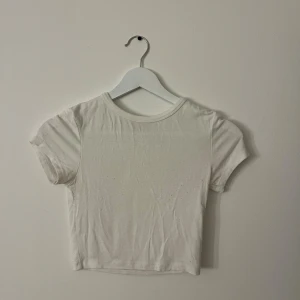 Vit croppad t-shirt i bomull - Säljer en enkel och stilren vit croppad t-shirt med rund hals och korta ärmar. Toppen är i mjuk bomull och har en tight passform som framhäver figuren. Perfekt till jeans eller kjol för en clean look.