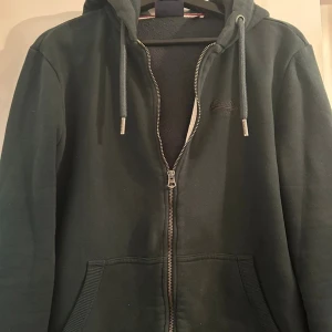 Mörkgrön hoodie från Superdry - Mörkgrön hoodie från Superdry med dragkedja, huva och snörning. Mjuk insida och två fickor framtill. Perfekt för chill dagar eller när du vill ha en avslappnad stil.