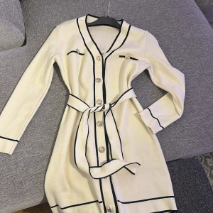 Vit stickad klänning med knappar från SHEIN - Supersnygg vit stickad klänning från SHEIN med svarta detaljer, guldiga knappar och bälte i midjan. Långärmad och med markerade svarta kanter för en chic look. Perfekt för dig som gillar klassisk stil med en twist.
