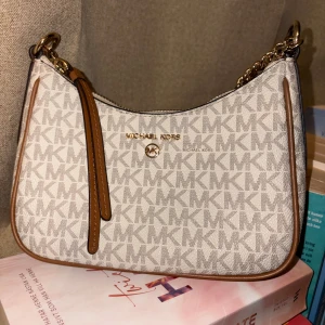 Michael Kors axelväska beige/brun - Snygg axelväska från Michael Kors i beige med bruna detaljer och guldiga kedjor. Väskan har MK-logga över hela ytan, dragkedja upptill och flera fack inuti. Perfekt storlek för det viktigaste och ger en lyxig känsla till din outfit.