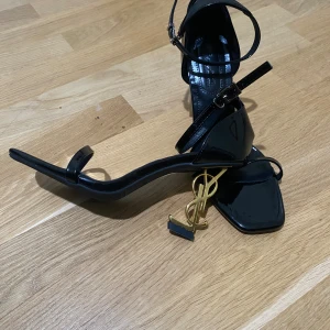 Svarta YSL Klacka med guld färg logga  - Säljer ett par svarta klackor från Saint Laurent med ikonisk YSL-logga i guld som klack. Perfekta för dig som vill sticka ut med en lyxig och edgy vibe.