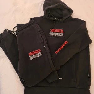 Svart dress från Hoodrich med tryck - Svart hoodie och byxa från Hoodrich med röd och vit logga på bröstet och ärmen. Tröjan har huva med snörning och stor magficka framtill. Mjuk insida och klassisk streetwear-look, perfekt för dig som gillar stilrena och bekväma plagg. 