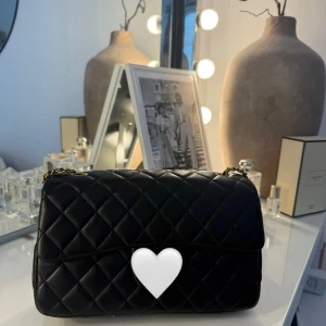 Svart quiltad axelväska med kedja - Stilren svart axelväska i quiltat skinn med klassisk kedjerem i guld och svart. Väskan har lockstängning och ett extra fack på baksidan. Perfekt för dig som gillar tidlös design och vill ha en väska som sticker ut med sin lyxiga känsla.