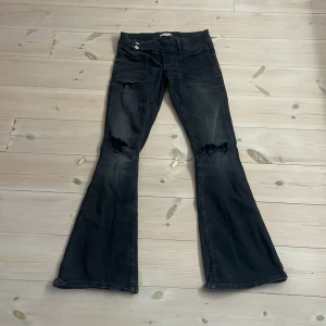 Svarta bootcut jeans från Gina Tricot - Säljer ett par svarta bootcut jeans från Gina Tricot med slitna hål på knäna och cool tvätt. Jeansen har låg midja, två knappar i linningen och stora fickor fram. Perfekta för dig som gillar en edgy och trendig look.