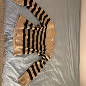 Randig stickad tröja Holly & Whyte - Beige och svart randig stickad tröja från Holly & Whyte. Tröjan har långa ärmar, ribbade muddar och fyra snygga guldknappar på ena axeln som detalj. lite nopprig och litet hål lvid handen men inget man märker av! (priset är som de är pga att den är nopprig och använd sen innan)