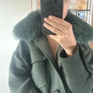 Grön kappa med päls från Meotine - Säljer en mörkgrön kappa från Meotine med stor fluffig päls runt kragen. Jackan har en klassisk rak passform, stora fickor framtill och stängs med knappar. Perfekt statement-plagg för kyliga dagar.
