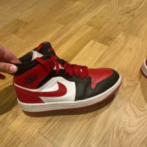 Nike Air Jordan 1 Mid Chicago - Säljer ett par Nike Air Jordan 1 Mid Chicago med klassisk färgblockning i rött, svart och vitt. Skorna har röd swoosh, svarta och röda paneler, vita partier och röda skosnören. Tillverkade i skinn med perforerad tå och ikonisk Air Jordan-logga på sidan.