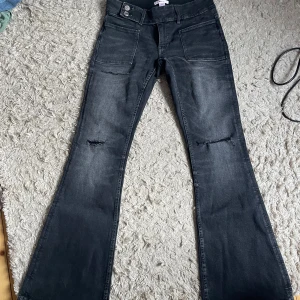 Svarta bootcut jeans från Gina Tricot - Snygga svarta jeans från Gina Tricot i bootcut-modell med slitningar på knäna och stora fickor framtill. Jeansen har en lätt tvättad look och dubbla knappar i midjan. Perfekta för dig som gillar en trendig och avslappnad stil.