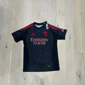 Svart Benfica fotbollströja Adidas - Snygg svart Benfica fotbollströja från Adidas med röda detaljer och tryck. Tröjan har korta ärmar, röda ränder på axlarna och subtilt mönster över hela framsidan. Materialet är lätt och andas, perfekt för träning eller match.