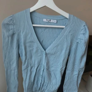 Ljusblå kofta från NA-KD - Supersöt ljusblå kofta från NA-KD med v-ringning och knappar framtill. Aldrig använd!