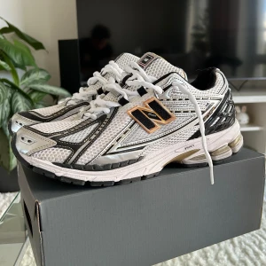 New Balance 1906R vita sneakers - Säljer ett par New Balance 1906R sneakers i vitt, svart och silver med guldiga detaljer. Skorna har mesh-ovandel, svarta och vita detaljer samt en chunky sula med stötdämpning. Klassisk N-logga på sidan och snörning framtill. Perfekta för en sportig och trendig look. Använda typ 2-3 gånger helt nya. Storlek 45 men sitter som 43-44