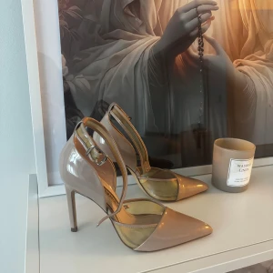 Beige pumps med spetsig tå och klack - Säljer ett par stilrena beige pumps med spetsig tå och hög klack. Skorna har en elegant rem runt ankeln och är tillverkade i glansigt syntetmaterial. Perfekta för dig som vill ha en chic och modern look.