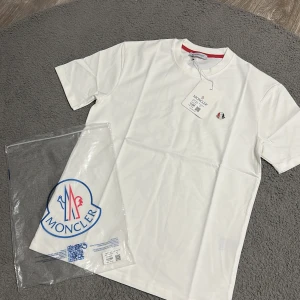 Vit Moncler t-shirt med logga - Vit t-shirt från Moncler i mjuk bomull med klassisk rund hals och korta ärmar. Diskret Moncler-logga på bröstet och röd detalj på insidan av kragen. Perfekt för dig som gillar stilrena och exklusiva plagg.