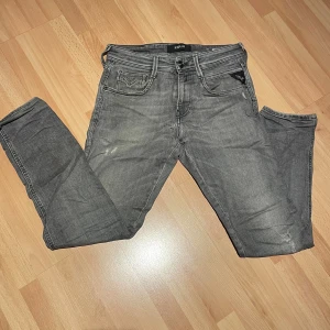 Replay Anbass grå jeans, stl 29 - Säljer ett par Replay Anbass jeans i grå tvätt med klassisk femficksdesign. Jeansen har diskreta slitningar och snygga detaljer på bakfickorna. Perfekta för dig som gillar en stilren och avslappnad look. Det är ett litet hål i skrevet men det är inte stort och lätt att sy för bara syra jag inte orkar göra det.