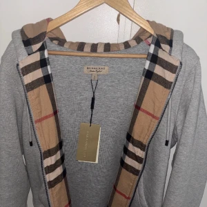Grå Burberry hoodie med klassiskt mönster - Grå hoodie från Burberry med dragkedja och huva. Insidan av huvan har det ikoniska rutiga Burberry-mönstret i beige, svart och rött. Broderad logga på bröstet. Perfekt för dig som vill ha en stilren och lyxig vardagslook.