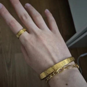 Guldigt armband och ring med romerska siffror - Säljer ett matchande set med armband och ring i guldig metall. Båda har ingraverade romerska siffror runt om för en stilren och modern look. Perfekt för dig som gillar accessoarer med unika detaljer och vill sticka ut lite extra.