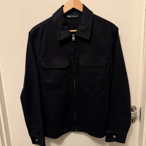 Svart overshirt från Zara, storlek M - Dom vita prickarna är inte stains är bara nåt vit som kom på tröjan när jag la den på golvet! Säljer en svart overshirt från Zara i storlek M. Jackan har raka linjer, klassisk krage, två bröstfickor med lock och dragkedja framtill. Materialet är bomull och modellen är långärmad med knappdetaljer vid ärmslut. Perfekt för lager-på-lager och stilren look.