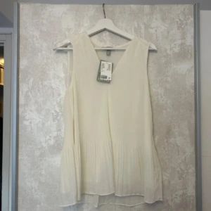 Vit ärmlös blus från H&M - Säljer en luftig vit blus från H&M i storlek S. Blusen är ärmlös med v-ringning och har ett lätt plisserat tyg som ger en flowy känsla. Perfekt för varma dagar och enkel att styla med jeans eller kjol.