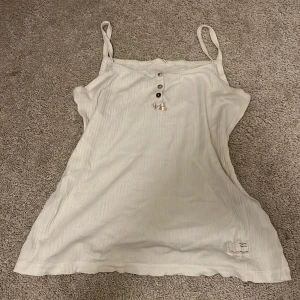 Vit ribbad linnetopp med knappar - Supersöt vit ribbad linnetopp från Odd Molly med smala axelband, tre knappar framtill och en liten rosett. Broderad detalj på ryggen med hjärtan och krona. Lätt och luftig, perfekt för varma dagar.