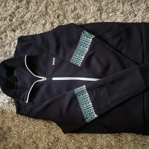 Svart zipjacka från BOSS, storlek S - Svart jacka från BOSS med hel dragkedja och vit piping längs kragen. Tryckt BOSS-logga på bröstet och upprepade loggor i turkos och vitt på ärmarna. Mjuk och sportig känsla, perfekt för en chill look.