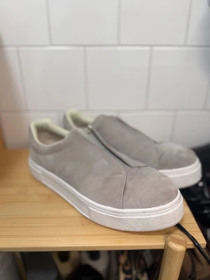 Eytys sneakers  - Snygga slip-on sneakers från Eytys i ljusgrå mocka med vit sula och lila detaljband bak. Skorna har rund tå och minimalistisk design utan snörning. Insidan har korksula och krämvit kant. Perfekta för dig som gillar stilrena och bekväma skor. Storlek: 45