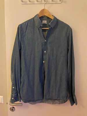 Blå skjorta från Filippa K i bomull (M) - Snygg blå skjorta från Filippa K i storlek medium. Denimfärg. Skjortan har klassisk krage, långa ärmar med manschett och vita knappar framtill. Materialet är mjukt och 100% bomull och passformen är normal. Perfekt för dig som gillar stilrena och enkla plagg.