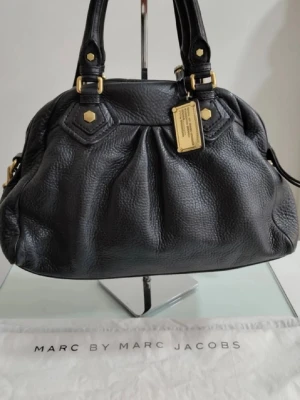 Svart handväska från Marc Jacobs - Säljer en snygg svart handväska från Marc by Marc Jacobs i mjukt skinn med guldfärgade metalldetaljer och dekorativ bricka framtill. Väskan har dubbla handtag och en klassisk, rundad form som ger en cool och tidlös vibe.