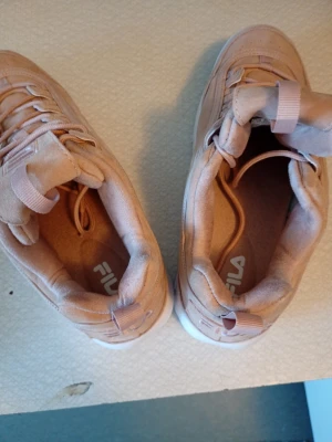 Beige sneakers från Fila i mocka - Säljer ett par snygga beige sneakers från Fila med ovandel i mjuk mocka och vita sulor. Skorna har rund tå, snörning och diskreta detaljer som ger en clean look. Perfekta till jeans eller kjol för en chill vardagsstil.