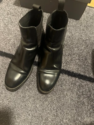 Svarta chelsea boots i läder stl 40  - Säljer ett par svarta chelsea boots med rund tå och platt sula. Skorna är tillverkade i slätt skinn och har elastiska paneler på sidorna samt en praktisk flik baktill för enkel påtagning. Perfekta för dig som gillar stilrena och klassiska boots. Fint begagnat skick :) 