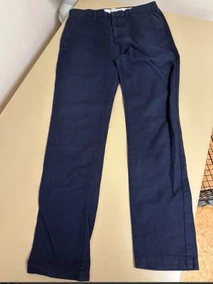 Mörkblå chinos från Jack & Jones W32 L34 slim fit  - Snygga mörkblå chinos från Jack & Jones i storlek W32 L34. Tillverkade i mjuk bomull med lite stretch för extra komfort. Klassisk rak passform med bälteshällor och sidofickor. Perfekta till en avslappnad men ändå stilren look.Har ett litet hål vid ena fickan, i övrigt inget att anmärka på. Fint begagnat skick.