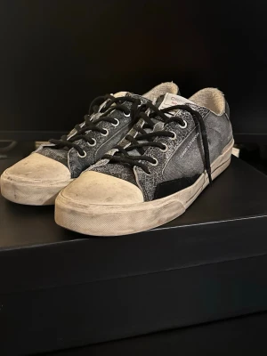 Svarta sneakers från Crime London - Säljer ett par svarta sneakers från Crime London med slitna detaljer och vita sulor. Skorna har snörning, metallnitar bak och textilmaterial med inslag av skinn. Märkeslogga på sidan och hälen. Perfekta för dig som gillar en rå och urban stil.