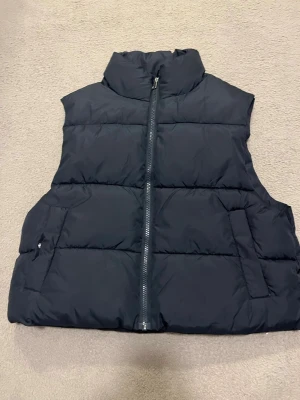 Svart dunväst från 157, storlek M - Säljer en svart dunväst från 157 i storlek M. Västen har hög krage, raka kanter och en rejäl dragkedja framtill. Den är quiltad och har två sidofickor. Perfekt för lager-på-lager under kyliga dagar. Enkel och stilren design som funkar till många outfits.