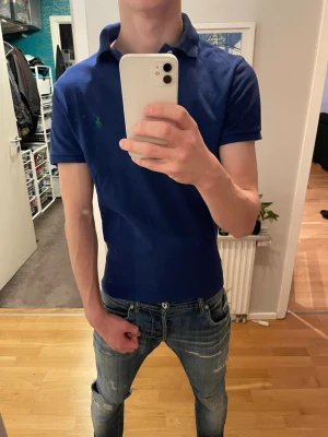 Marinblå pikétröja Ralph Lauren S - Klassisk marinblå pikétröja från Ralph Lauren i slim fit. Tröjan har korta ärmar, krage med två knappar och den ikoniska gröna broderade loggan på bröstet. Tillverkad i 100% bomull för en skön och luftig känsla.