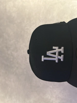 Svart LA 59FIFTY keps från New Era - Säljer en svart LA-keps från New Era, modell 59FIFTY. Kepsen har broderad LA-logga i vitt framtill, MLB-logga bak och New Era-logga på sidan. Klassisk flat peak och stilren look, perfekt för dig som gillar streetwear. Har aldrig använt den köpte den i somras då den var för stor sedan tänkte jag att jag kanske växer i den men det gör jag inte så säljer den nu.