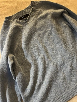 Blå stickad tröja från Vero Moda - Mysig blå stickad tröja från Vero Moda med rund hals och ribbade muddar vid hals, ärmslut och nederkant. Perfekt för kyliga dagar och enkel att matcha med jeans eller kjol. Tröjan har en klassisk, loose passform och långa ärmar.