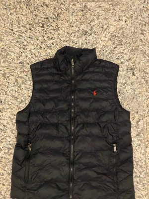 Svart dunväst från Polo Ralph Lauren - Säljer en svart dunväst från Polo Ralph Lauren med klassisk quiltad design och dragkedja framtill. Västen har två fickor med dragkedja och en röd Polo-logga på bröstet. Perfekt för lager-på-lager under höst och vinter.