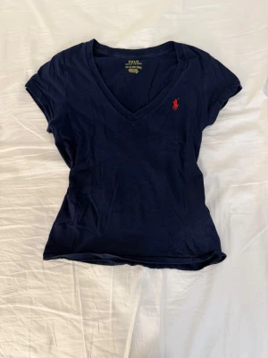 Mörkblå v-ringad t-shirt Polo Ralph Lauren - Snygg mörkblå t-shirt från Polo Ralph Lauren med klassisk röd broderad logga på bröstet. T-shirten har v-ringning och korta ärmar, tillverkad i mjuk bomull för skön känsla. Perfekt till jeans eller shorts för en clean och enkel look.