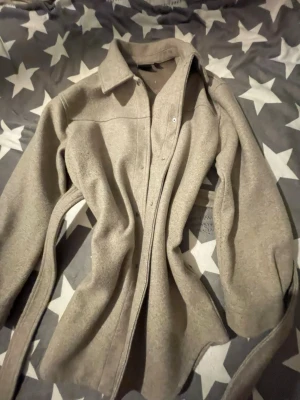 Beige kappa från Vero Moda - Säljer en kappa overshirt från Vero Moda i storlek M. Jackan har klassisk krage, långa ärmar och knappar framtill. Materialet är mjukt och känns fleece-liknande, perfekt för lager på lager. Enkel och stilren design som funkar till många outfits.