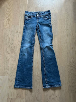 Gina Tricot (146) - Blå bootcut jeans med låg midja - Snygga blå jeans med bootcut-snitt och låg midja. Klassisk femficksmodell med kontrastsömmar och slitningar framtill. Jeansen är tillverkade i ett mjukt bomullsmaterial och har bälteshällor samt knapp- och dragkedjestängning. Perfekta för en chill och avslappnad stil.