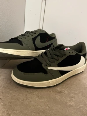 Nike Air Jordan 1 Low Olive/Black - Nike Air Jordan 1 Low sneakers i olivgrönt och svart med vita swoosh-loggor och röda broderade detaljer på hälen. Skorna har snörning, platt sula och rund tå. Materialet är en mix av skinn och syntet, med klassisk Air Jordan branding på baksidan och yttersulan i olivgrönt.