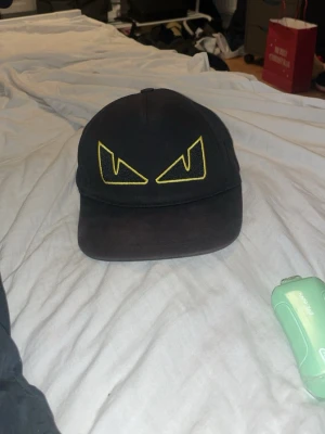 Svart keps med gula ögon - Säljer en svart keps med justerbart spänne bak och broderade gula ögon framtill. Kepsen har klassisk form och är tillverkad i bomullstyg. Perfekt för dig som vill sticka ut med en cool detalj på huvudet.