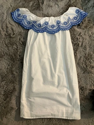 Vit offshoulder klänning med blå broderi - En vit kort klänning med offshoulder och fin blå broderad spets upptill. Perfekt till sommarn! Nästan aldrig använd, bara någon gång.