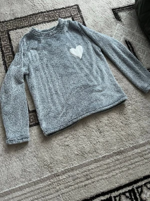 Grå fluffig tröja med hjärtdetalj - Supermjuk och mysig grå tröja från Cubus i storlek XS. Tröjan är gjord i fluffigt fleece-material och har en vit hjärtapplikation på bröstet. Perfekt för kalla dagar och har långa ärmar samt rund hals.