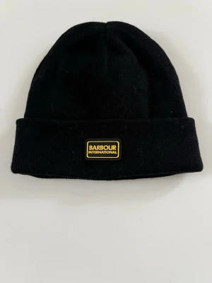 Svart mössa från Barbour International - Klassisk svart mössa från Barbour International med uppvikt kant och gul logotyp framtill. Tillverkad i mjuk ullblandning som håller dig varm och stilren under kyliga dagar. Enkel och clean design som passar till det mesta.
