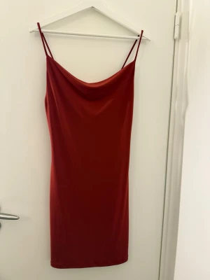 Röd axelbandslös kortklänning - Säljer en snygg röd klänning med tunna axelband och draperad halsringning. Klänningen är kort och har en slät, figurnära passform. Materialet känns mjukt och har ett lätt glansigt utseende, perfekt för dig som gillar minimalistisk stil.