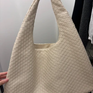 Beige axelväska med flätad struktur - Säljer en stilren beige axelväska med unik flätad struktur. Väskan har en mjuk och flexibel form som gör den enkel att bära över axeln. Perfekt för dig som gillar minimalistisk design och vill ha en väska som sticker ut lite extra.