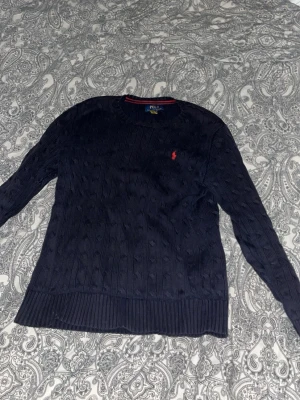 Mörkblå stickad tröja från Polo Ralph Lauren - Snygg mörkblå stickad tröja från Polo Ralph Lauren med klassiskt kabelstickat mönster och röd broderad logga på bröstet. Tröjan har rund hals, långa ärmar och ribbade muddar vid ärmslut och nederkant. Perfekt för dig som gillar stilrena och tidlösa plagg.