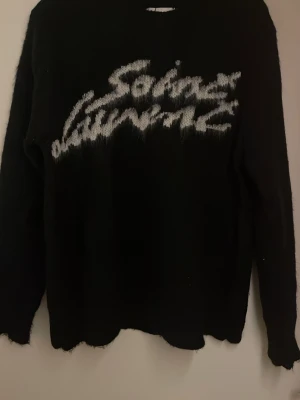 Svart stickad Saint Laurent tröja - Cool svart stickad tröja från Saint Laurent med vit logotyp framtill i en grafisk stil. Tröjan har lång ärm och en lite ruffad kant nedtill för en edgy vibe. Perfekt för dig som gillar designer och vill sticka ut med en statement piece.